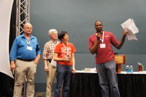 Audience members critiquing FOSS artifacts at POSSCON 2011; the image shows several people at the front of a room, with one man holding up a sheet of paper he is talking about into a microphone.