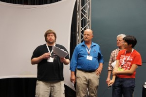 Audience members critiquing FOSS artifacts at POSSCON 2011; the image shows several people listening to a student in a black shirt explaining the webpage his group reviewed.